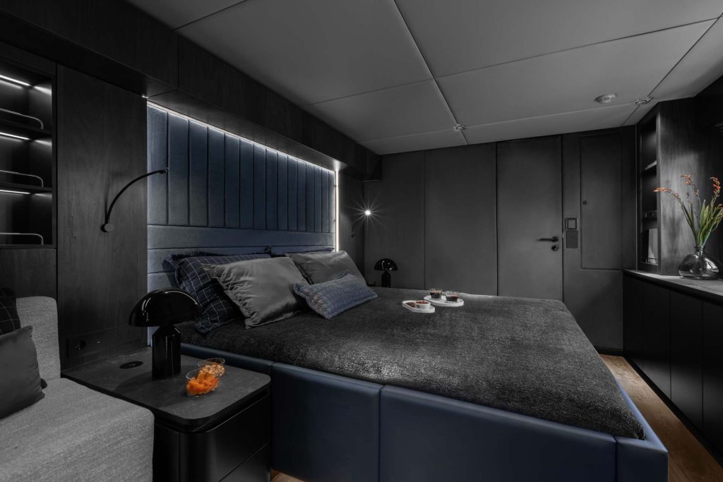 Interior view of a luxurious bedroom on a Sunreef Yachts vessel, featuring a dark, modern design with a large bed, seating area, and ambient lighting.